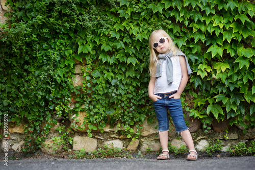 Funny little girl wearing sunglasses posing by bindweed wall on warm and sunny summer day