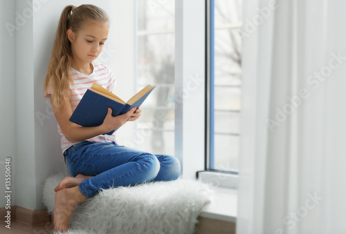 Cute little girl with book sitting on windowsill at home