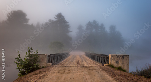 Low cloud and darkened skies.  Single point perspective, surreal coloured subdued light from summer storm over a single lane, steel & timber bridge spanning a narrow passage in a lake.