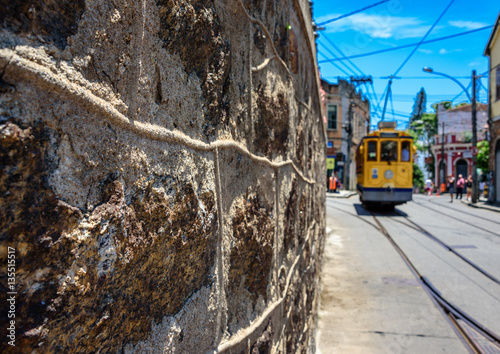 The stone wall, blue sky and old-fashioned yellow tram bonde going by tram tracks in Santa Teresa district of Rio de Janeiro, Brazil