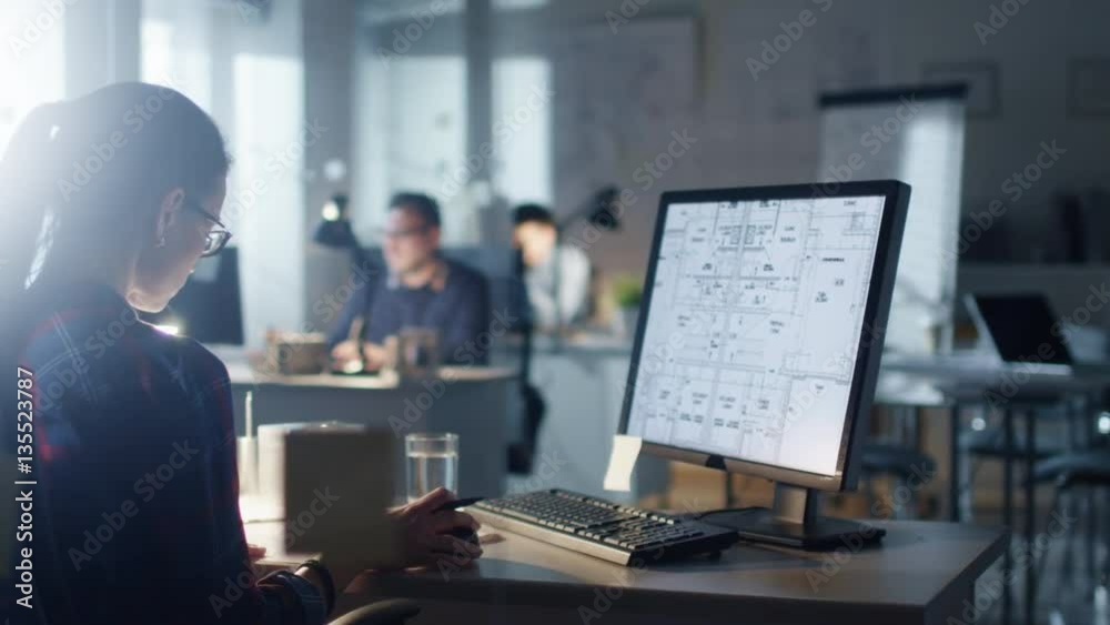 Late at Night in the Engineering Bureau Female Designer Works on a Personal Computer that Shows Blueprints of Her Project. In the Background Her Colleagues Working. Shot on RED Cinema Camera 4K (UHD).