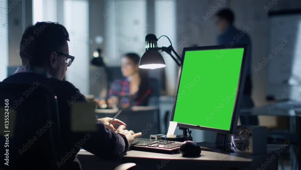 Late at Night  Industrial Engineer Sits at His Desk Working on a Computer with Green Screen on. Office is Modern. In the Background Colleagues are Working at Their Desks.  

