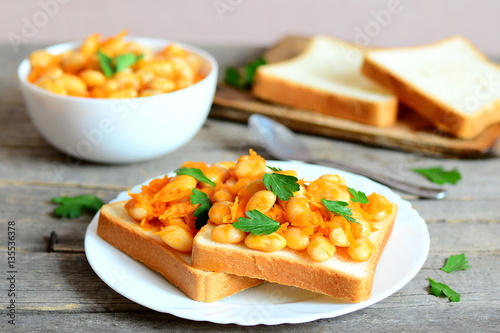 Open sandwiches with baked white beans. Beans baked with carrots, garlic and tomato sauce on a bowl, bread slices, spoon, fresh parsley on a rustic wooden table. Vegetarian and vegan protein dish