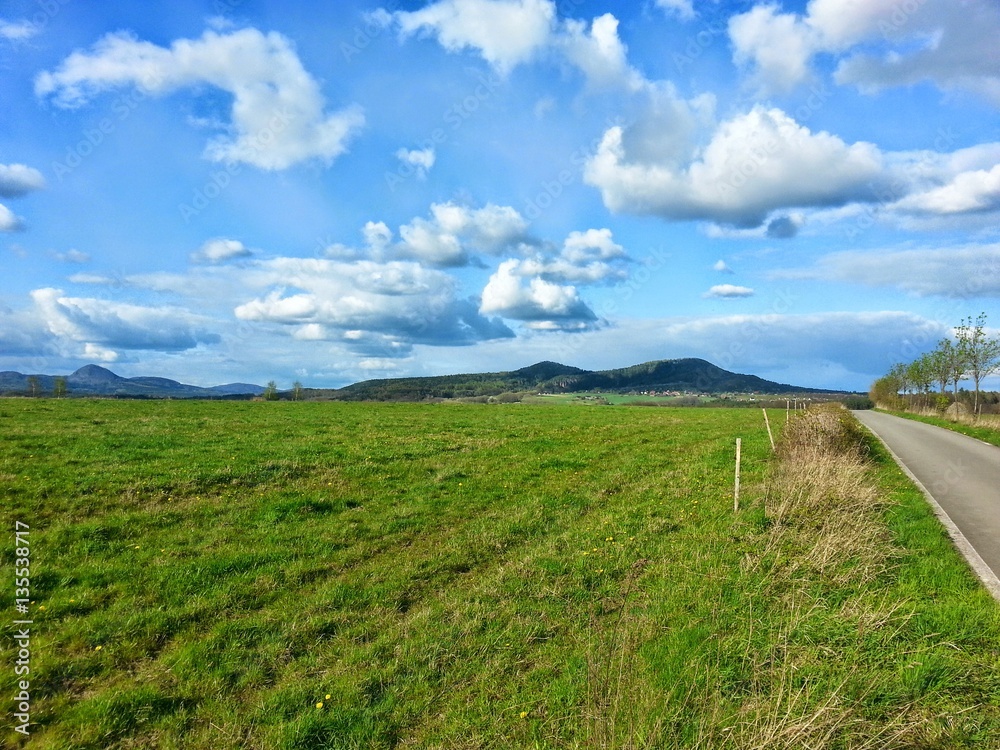 Obraz premium The blooming meadow with nice blue sky with dispersed clouds by sunny day. The Czech nature. Hills in the background.