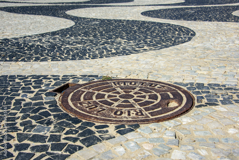 Black and white iconic mosaic, Portuguese pavement by old design pattern with a manhole at Copacabana Beach, Rio de Janeiro, Brazil Black and white iconic mosaic, Portuguese pavement by old design pattern with a manhole at Copacabana Beach, Rio de Janeiro, Brazil