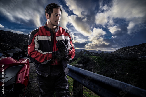 Male motorcyclist wearing protective leather racing suit with a red bike or motorcycle on an open road.  The vehicle is cropped to become generic non branded. The image depicts travel and adventure. photo