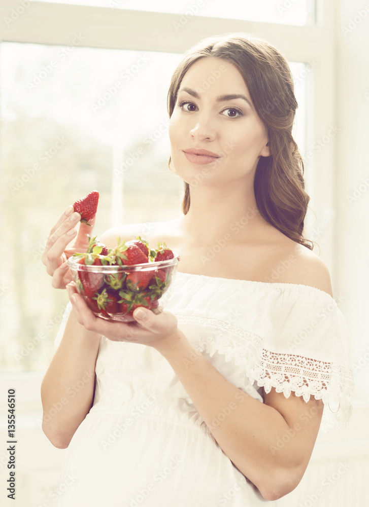 Beautiful pregnant woman eating strawberries in kitchen. Motherh Beautiful pregnant woman eating strawberries in kitchen. Motherh