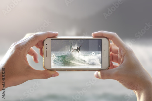 Photo camera of a smartphone. View through the screen the moment a young woman takes the picture. Surfer on a wave.