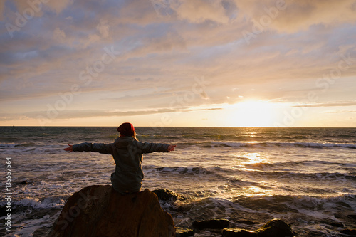 young woman sitting on a rock on the beach and looking at stormy sea and sunset sky. rear view.