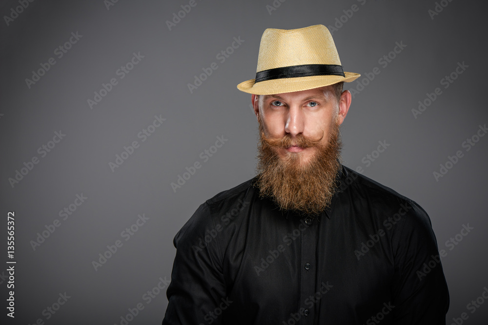 Closeup of hipster man with beard and mustashes wearing black shirt and straw fedora hat, over grey background Closeup of hipster man with beard and mustashes wearing black shirt and straw fedora hat, over grey background