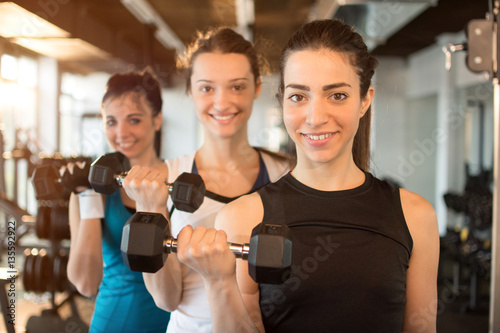 Three fit and beautiful young women lifting weights in a fitness club. Focus on the first girl in front.