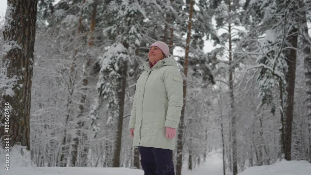 Elderly woman in warm clothing standing in the middle of snow-covered forest and admiring the beautiful scenery. Useful walking outdoors. Bright winter day.