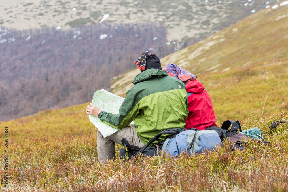 two tourists sitting on their back packs in mountains and exploring a travel map