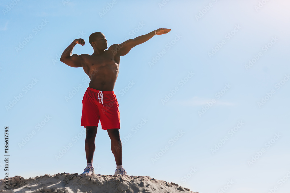 Strong Afro American man in red short shows muscles and looking upon a background blue sky Strong Afro American man in red short shows muscles and looking upon a background blue sky