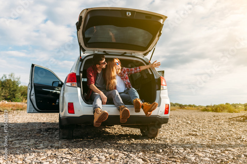 Happy traveler couple sitting in car open trunk and watch the sunrise