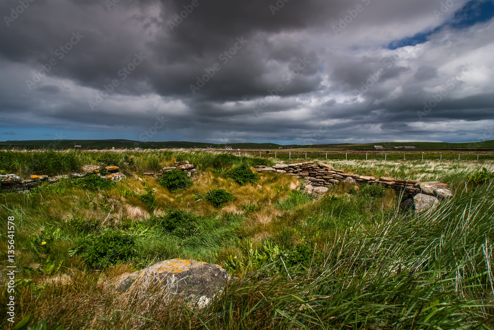 Orkneys, Scotland - June 5, 2012: Skara Brae Neolithic Settlement. Scary and vibrant scene of meadows, rocks and walls under a stormy, rainy, black sky with a thin blue line on the horizon.