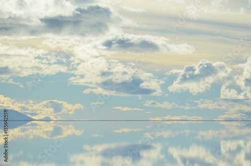 Céu azul e amarelado com nuvens baixas refletido por espelho natural de água no Salar de Uyuni