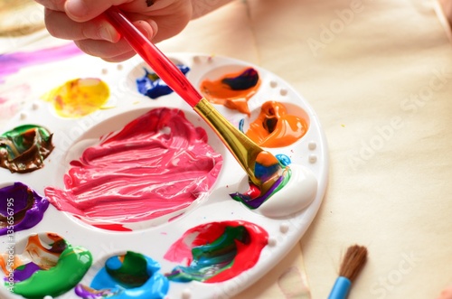Child's hand holding a paintbrush dipping into a palette full of colorful paint