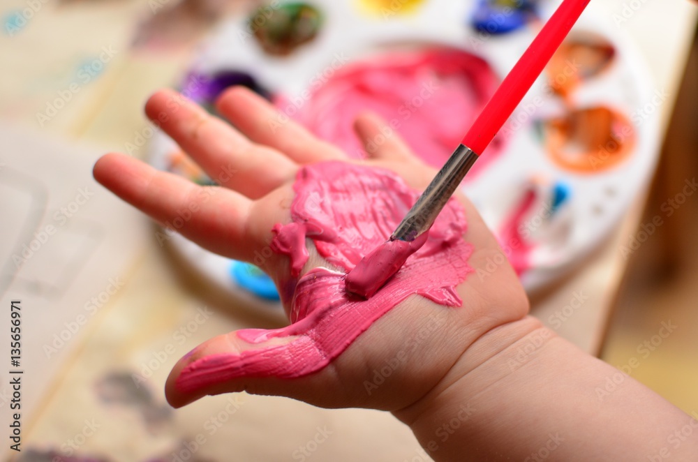 Fototapeta premium Child painting her hand with pink paint and background with palette of colorful paint