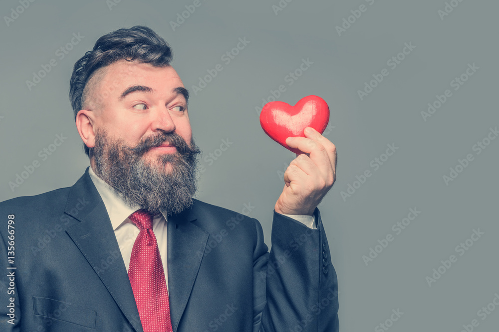 Smiling adult bearded man in a black suit and red tie holding a red heart. Gray background Smiling adult bearded man in a black suit and red tie holding a red heart. Gray background