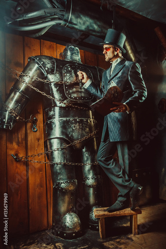 Young bearded man in cylinder hat is reading manual book while standing near huge robot.