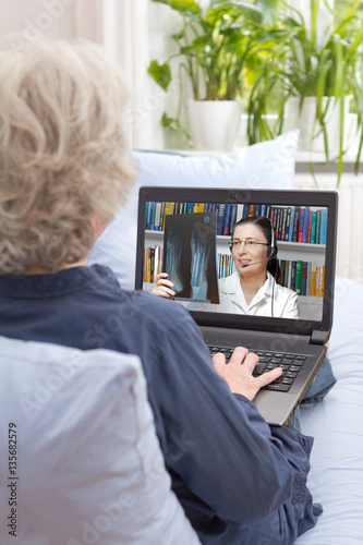 Rear view of a senior woman sitting with notebook on the couch of her living room, having a video call with her doctor via the internet, online consultation, e-health. photo