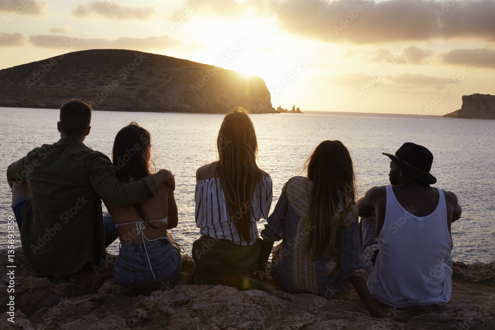 Rear View Of Friends Sitting On Cliff Watching Sunset Rear View Of Friends Sitting On Cliff Watching Sunset