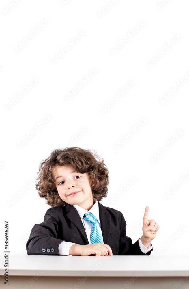 Little curly boy in a business suit sitting at a desk smiling out points with the index finger up. White background. Little curly boy in a business suit sitting at a desk smiling out points with the index finger up. White background.