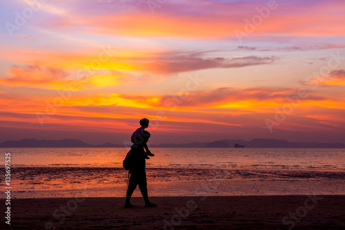 Father carrying his son on the neck walks on the beach at sunset over sea background. silhouette style