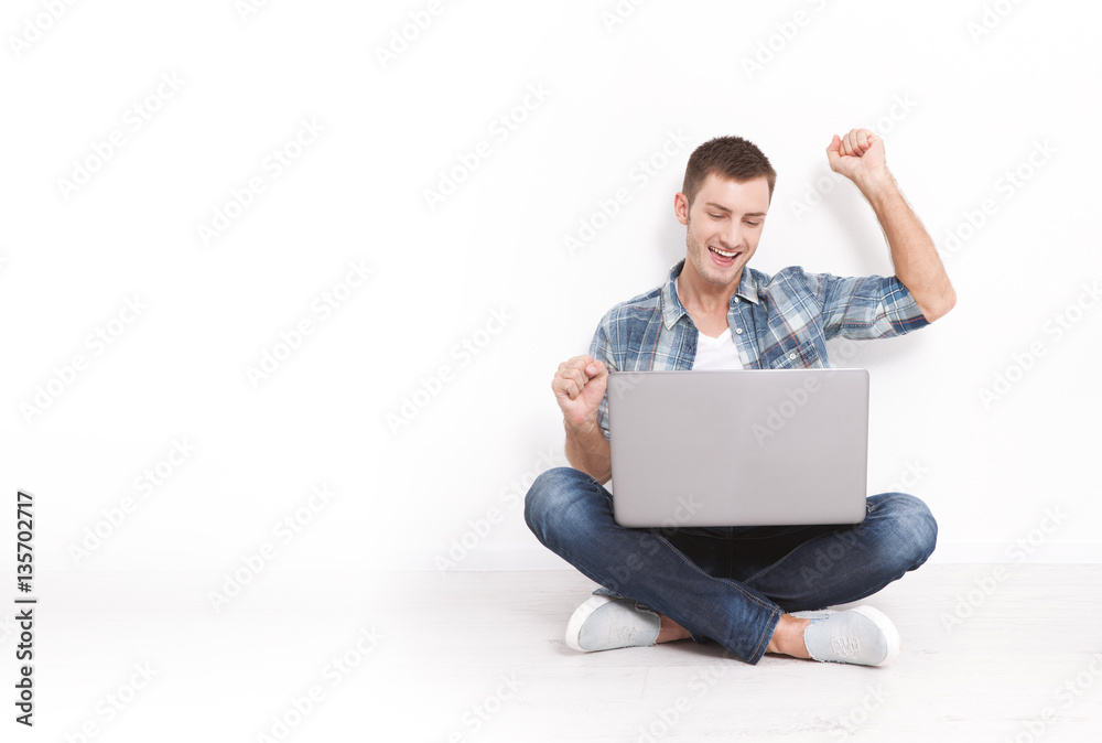 Handsome young man sitting on the floor near the wall, celebrating success with arms raised while looking at his laptop screen Handsome young man sitting on the floor near the wall, celebrating success with arms raised while looking at his laptop screen