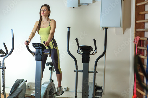 Young beautiful white girl in a yellow and gray sports suit is engaged on a stationary bike in the fitness club.