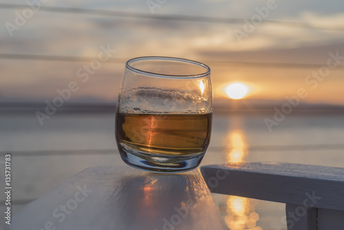 Single glass of whiskey on arm of deck chair at sunset