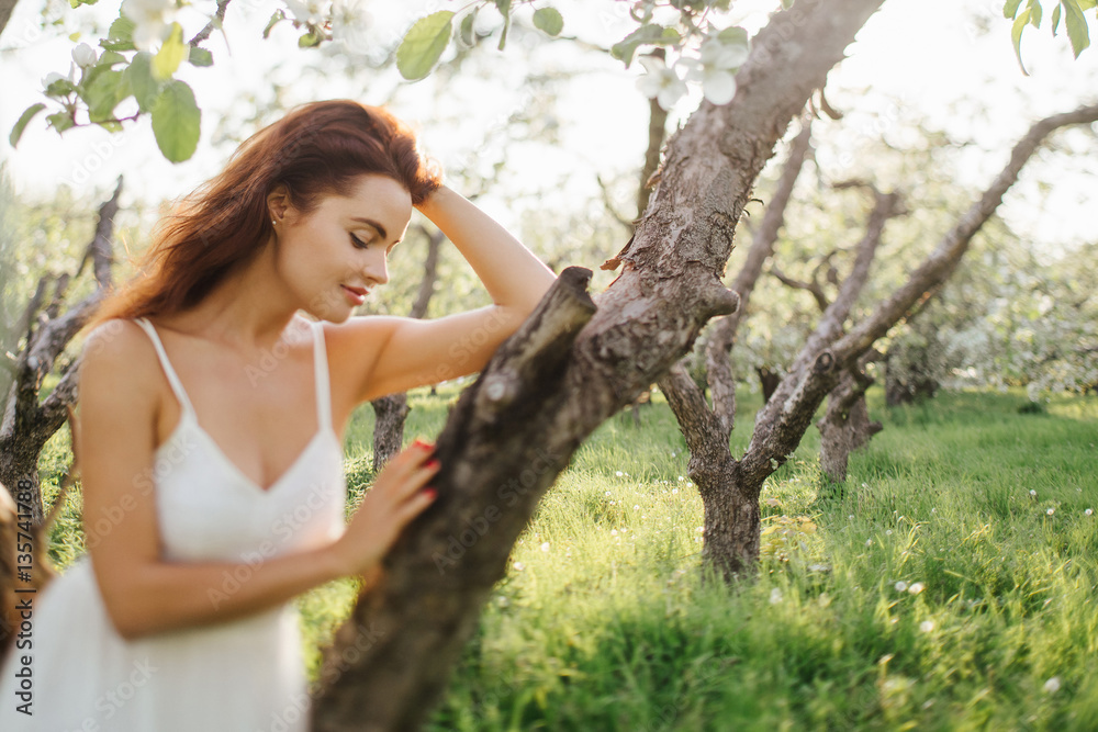 Young pretty serious caucasian woman in white dress standing near blooming spring tree. Youth, freshness, beauty, happiness concept. Tilt-shift image, intentionally blurred, soft selective focus.
