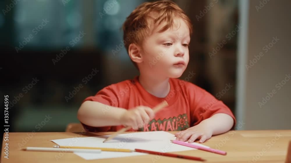 A cute little boy sitting at the table and drawing with the colored pencils on white paper. A cute little boy sitting at the table and drawing with the colored pencils on white paper.