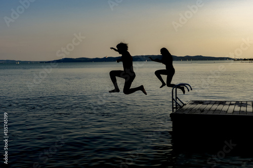 Shiluette of boy and girl jumping in the lake from pier