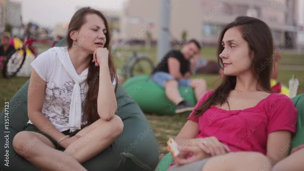 Two beautiful brunette girls sisters talking at the park. Sitting inbean bag chairs in summer day. Two beautiful brunette girls sisters talking at the park. Sitting inbean bag chairs in summer day.