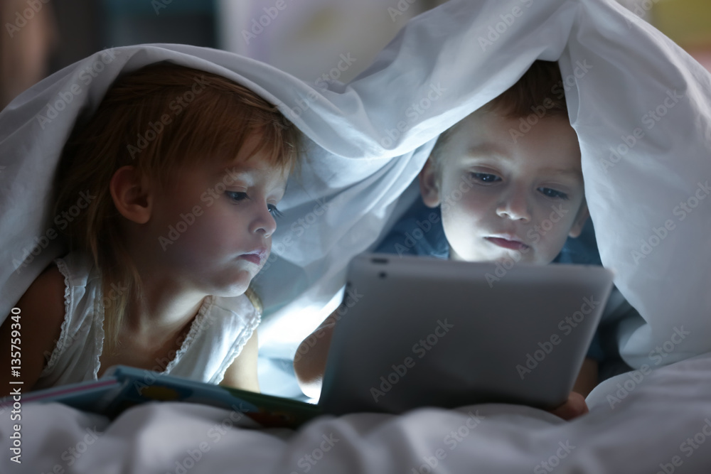 Cute little children lying under blanket with book and tablet computer