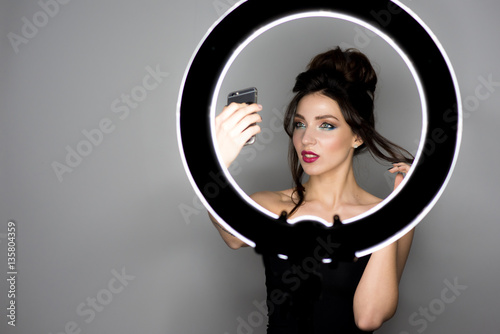 Studio shot of young and beautiful woman taking selfie picture with mobile phone in front of the ring lamp. Isolated on gray background. Place for text photo