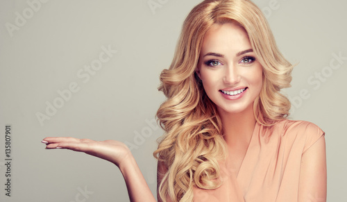 
    Blonde woman with curly hair shows your product . .Beautiful girl with beautiful smile pointing to the side . Presenting your product. Expressive facial expressions
