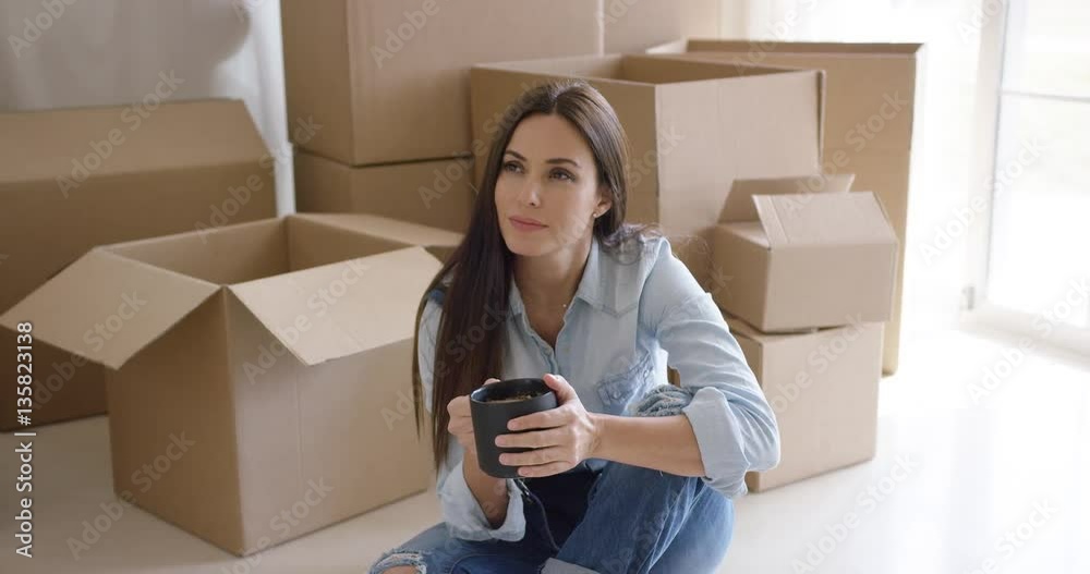 Thoughtful young woman squatting on the floor with a cup of coffee contemplating her new house surrounded by brown boxes Thoughtful young woman squatting on the floor with a cup of coffee contemplating her new house surrounded by brown boxes