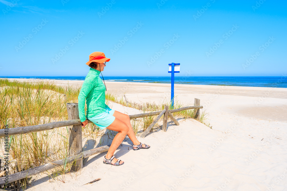Young woman sitting on wooden fence and looking at beautiful white sand beach, Baltic Sea, Poland