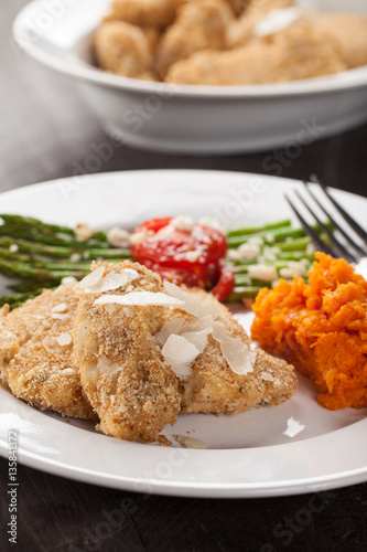 Parmesan Crusted Chicken Tenders with Roasted Sweet Potato and Roasted Asparagus topped with Campari tomatoes, Feta Cheese, Olive oil, and salt on a dark wooden background side view