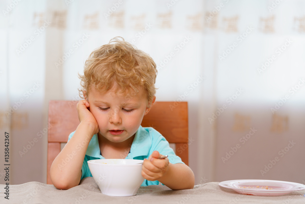 Blonde boy having breakfast while sitting on a chair at the kitchen table at home. White porcelain cup in front of kid. He thoughtfully looks inside it. Holds spoon with his hand. Blonde boy having breakfast while sitting on a chair at the kitchen table at home. White porcelain cup in front of kid. He thoughtfully looks inside it. Holds spoon with his hand.