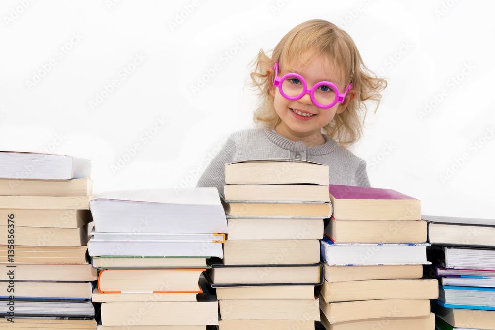 Three year old girl in pink glasses smiling over stack of  books. Isolated on white.