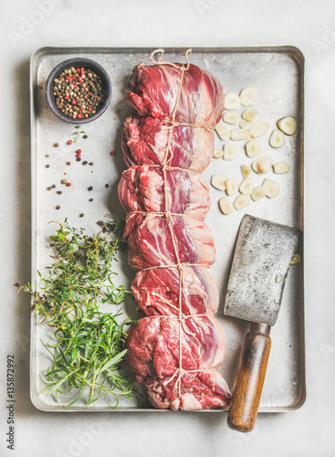 Raw uncooked roast beef meat cut with herbs, garlic and spices over light grey marble background, top view, vertical composition