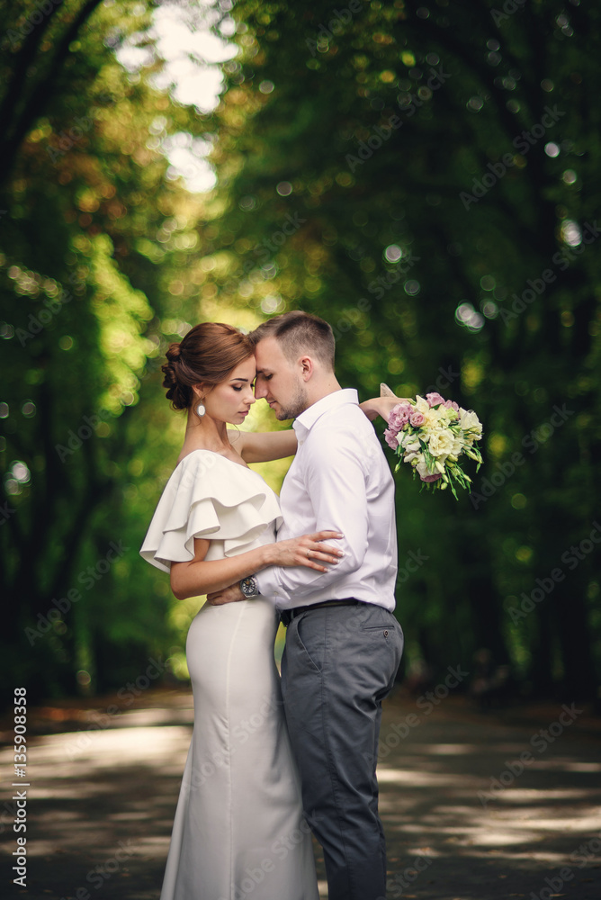 Handsome groom hugging his beautiful stunning bride with bouquet in romantic european park