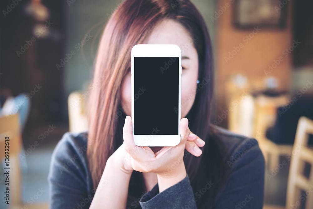 Mockup image of a beautiful woman holding and showing white mobile phone with blank black screen cover the face in modern cafe