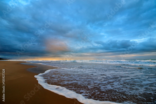 Seascape at sunset with waves on beach and heavy cloud cover.