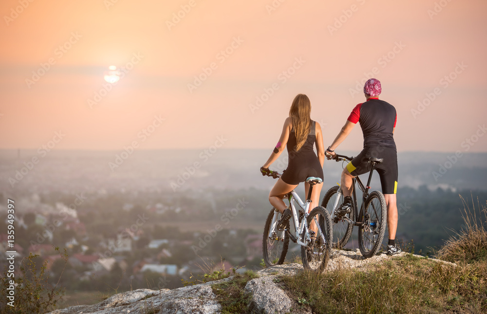 Rear view male and female bikers with moutains bicycles standing on the top of a hill enjoying the sunset. Blurred background with copy space