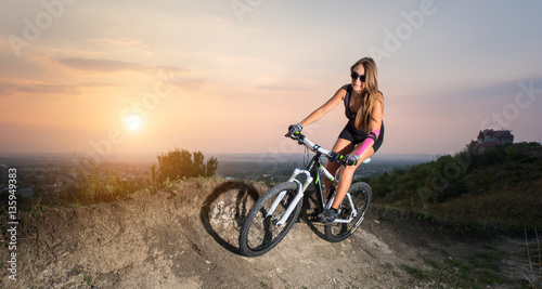 Fitness pretty girl in glasses riding on the mountain bicycle on a trail against evening sky with bright sun on the sunset. Pink Kinesio tape glued on the girl's hand.